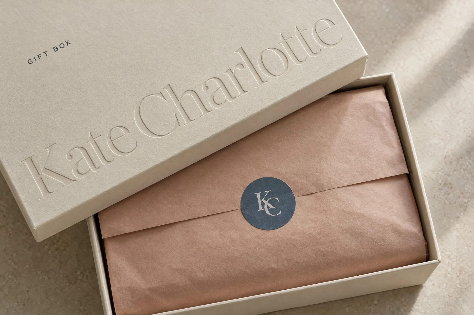 Ivory gift box with a blind-debossed wordmark, rose tissue and a slate KC seal